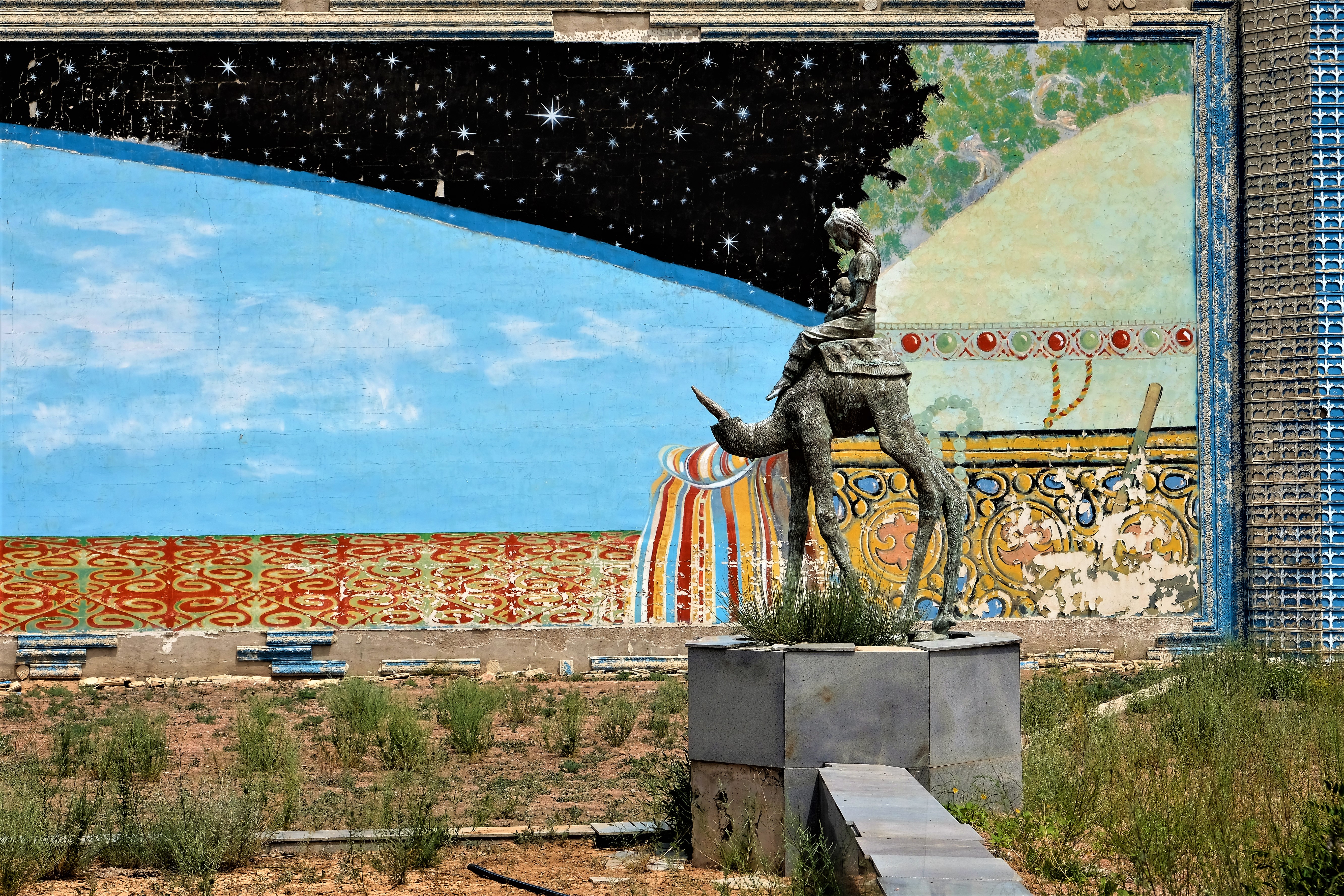 A statue of a girl riding a camel. The head of the camel is missing. A mural of the starry sky in the background