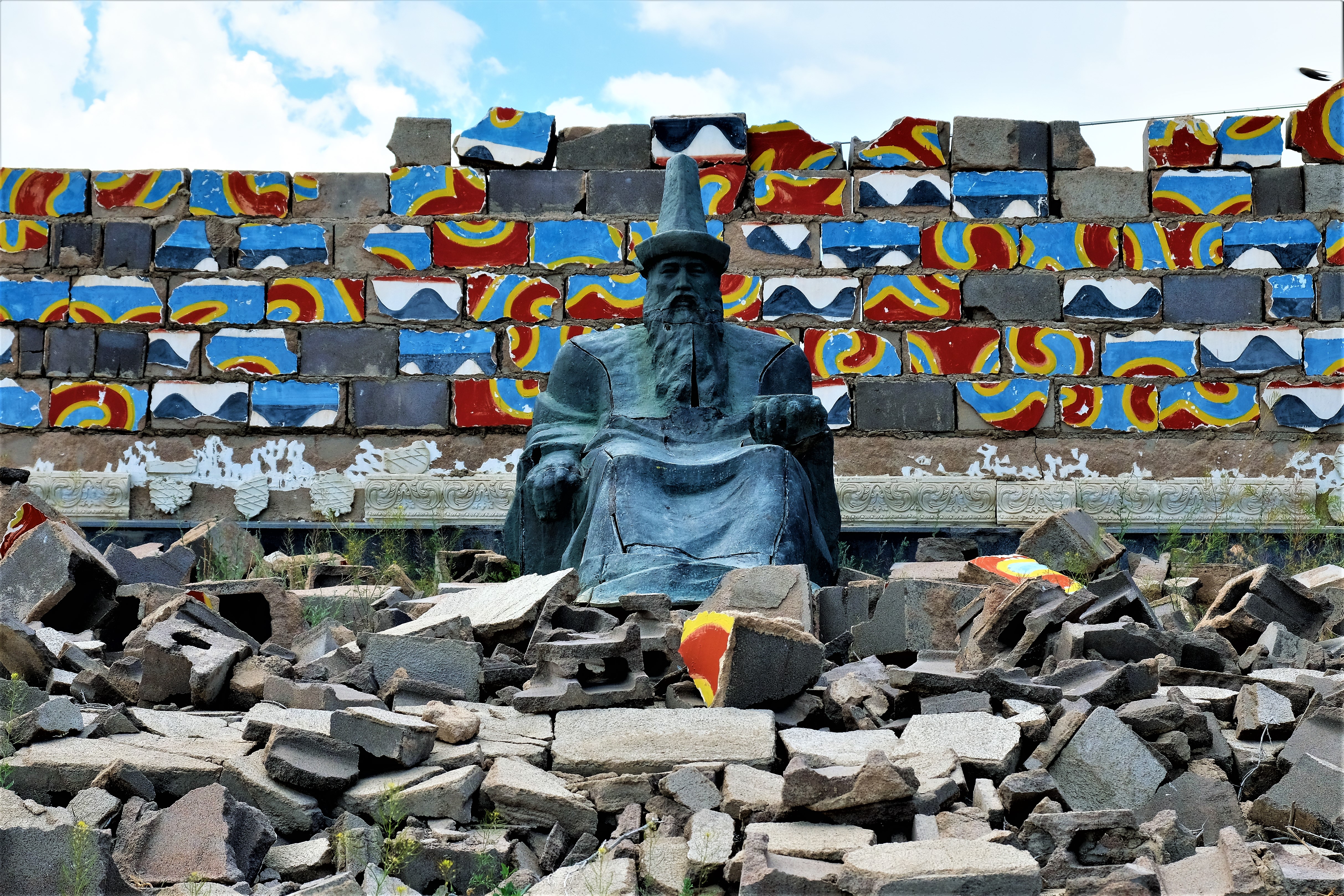 A statue of a sitting man in front of a trumbled wall