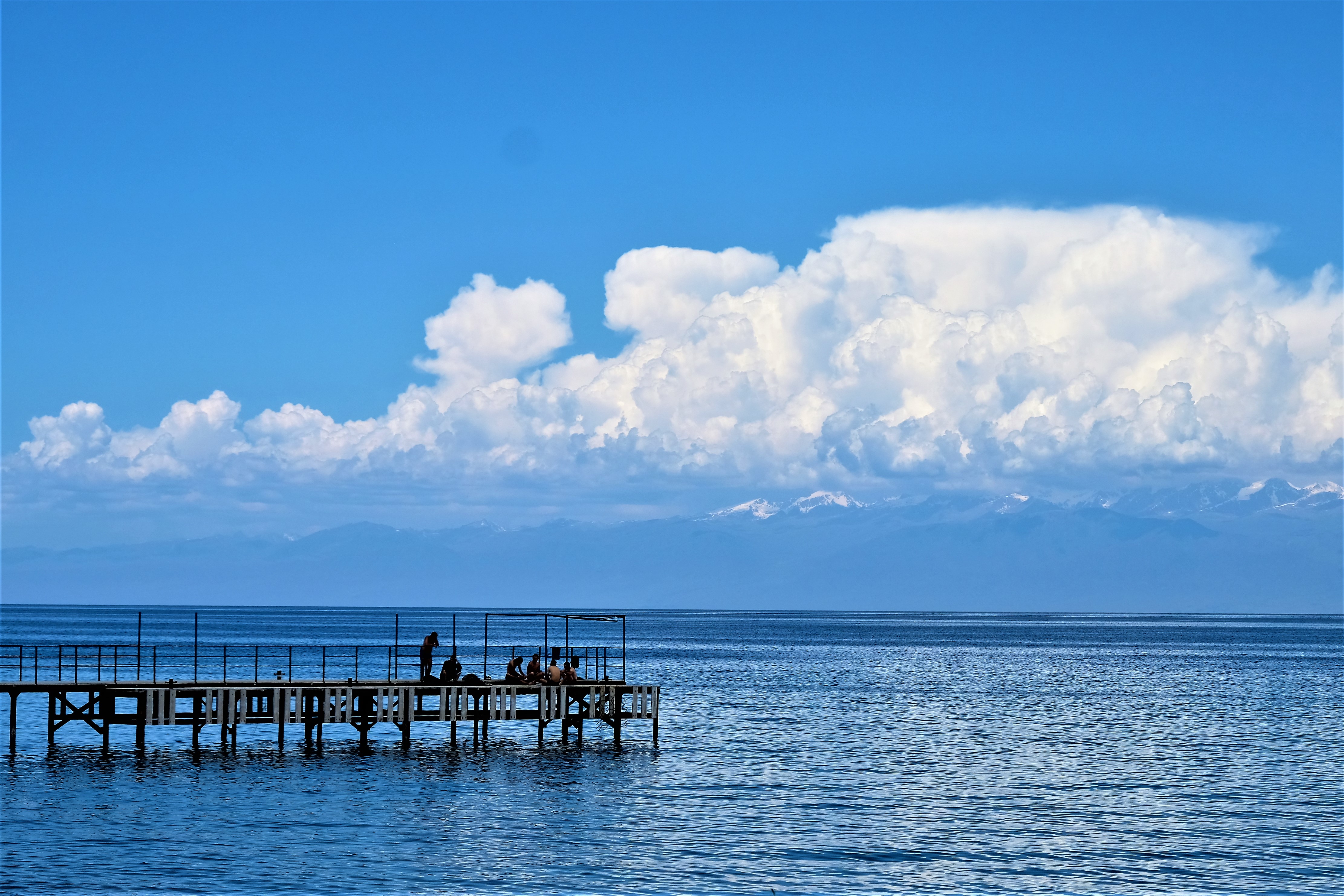 People sitting on a jetty at lake Issyk KUl in the sun, with big clouds and mountain range in the background